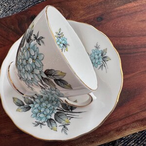 May include: A white teacup and saucer with a gold rim and a floral design featuring blue and green hydrangeas.