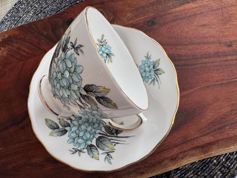 May include: A white teacup and saucer with a gold rim and a floral design featuring blue and green hydrangeas.