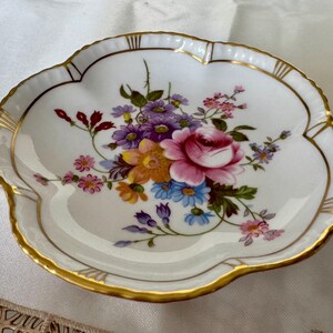 May include: A white, scalloped-edge dish with a floral bouquet design. The bouquet features a pink rose, purple, yellow, and blue flowers. The rim is trimmed with gold. A decorative piece for serving or display.