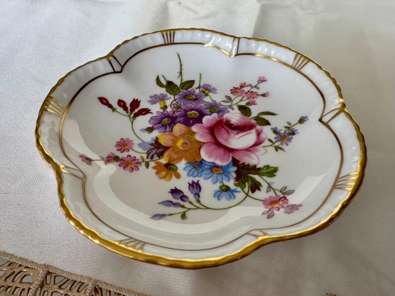 May include: A white, scalloped-edge dish with a floral bouquet design. The bouquet features a pink rose, purple, yellow, and blue flowers. The rim is trimmed with gold. A decorative piece for serving or display.