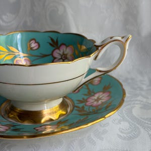 May include: A porcelain teacup and saucer set featuring a vibrant turquoise interior and border, decorated with pink floral motifs and gold leaf accents. The cup has a scalloped rim and a gold-trimmed handle, resting on a matching saucer.