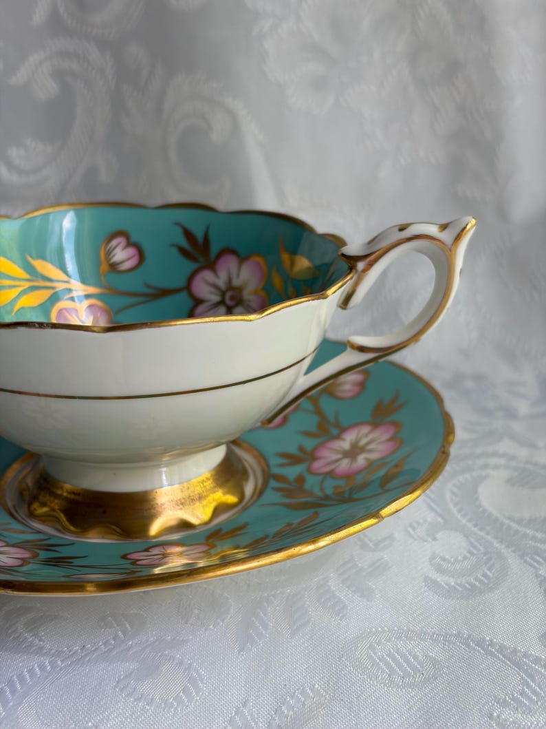 May include: A porcelain teacup and saucer set featuring a vibrant turquoise interior and border, decorated with pink floral motifs and gold leaf accents. The cup has a scalloped rim and a gold-trimmed handle, resting on a matching saucer.