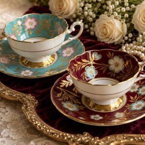 May include: Two porcelain teacups and saucers with scalloped edges. One set features a turquoise background with pink floral patterns and gold accents, while the other has a deep burgundy background with light blue floral patterns and gold trim.