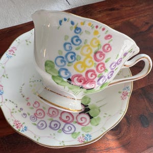 May include: A white teacup and saucer set with a floral design. The teacup has a colorful floral pattern with pink, yellow, blue, and green flowers. The saucer has a similar floral pattern with smaller flowers.