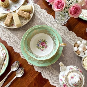 May include: A vintage tea setting with a mint green teacup and saucer, adorned with gold trim and a floral design. A tiered serving tray holds pastries and sandwiches. Pink roses and a teapot complete the elegant arrangement.