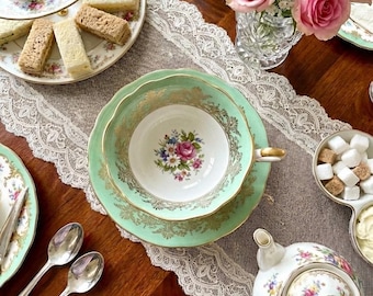 May include: A mint green teacup and saucer set with gold filigree trim and a central floral motif, accompanied by a white teapot with floral patterns, silver spoons, a tiered tray of pastries, finger sandwiches, and a vase of pink roses.