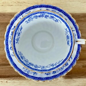 May include: A white teacup and saucer set with a blue floral pattern and gold trim. The teacup is round with a handle, and the saucer has a scalloped edge. The set is placed on a wooden surface.