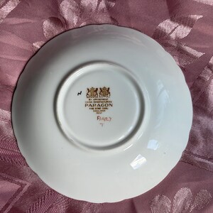 May include: A white porcelain saucer with a scalloped edge. The underside features a gold-colored maker's mark with the text BY APPOINTMENT CHINA MANUFACTURERS PARAGON FINE BONE CHINA ENGLAND REGD, along with a handwritten number and the letter T.