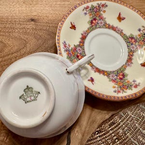 May include: A white teacup and saucer with a floral pattern. The saucer has a gold rim and the teacup has a gold rim and a gold crown with the words "Royal Ansley England" on the bottom.