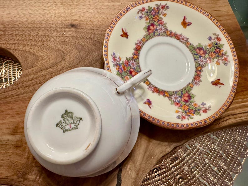 May include: A white teacup and saucer with a floral pattern. The saucer has a gold rim and the teacup has a gold rim and a gold crown with the words "Royal Ansley England" on the bottom.
