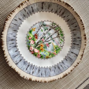 Pu&ograve; includere: Un piattino in porcellana bianca con un design a betulla grigio e oro. Il centro del piattino presenta una scena floreale colorata con un lago e alberi.