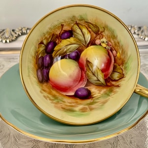 May include: A vintage teacup and saucer set. The teacup interior features a hand-painted still life of peaches, grapes, and leaves. The cup and saucer are light green with gold trim. The teacup has a gold handle.
