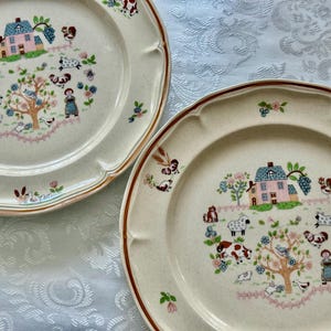 May include: Two off-white ceramic plates with a brown rim and scalloped edges. Each plate features a central design of a house, animals, and a tree with flowers. The plates have a rustic, farmhouse aesthetic.