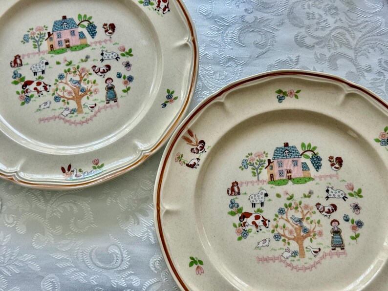 May include: Two off-white ceramic plates with a brown rim and scalloped edges. Each plate features a central design of a house, animals, and a tree with flowers. The plates have a rustic, farmhouse aesthetic.
