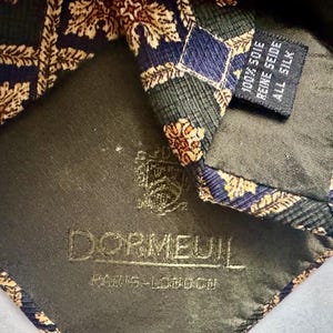 May include: A necktie featuring a dark blue and green diamond pattern with gold-toned leaf motifs. The interior lining displays the embroidered text DORMEUIL PARIS-LONDON and a crest, with a tag stating 100% SOIE REINE SEIDE ALL SILK.