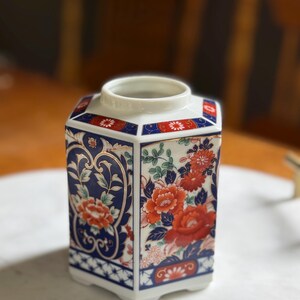 Peut inclure: Un vase hexagonal en c&eacute;ramique avec une base et un bord blancs. Le vase pr&eacute;sente un motif floral avec des fleurs rouges et oranges, des feuilles vertes et des accents bleus et dor&eacute;s. Le vase est probablement &agrave; des fins d&eacute;coratives.