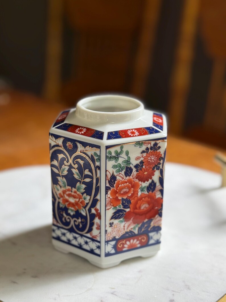 Peut inclure: Un vase hexagonal en c&eacute;ramique avec une base et un bord blancs. Le vase pr&eacute;sente un motif floral avec des fleurs rouges et oranges, des feuilles vertes et des accents bleus et dor&eacute;s. Le vase est probablement &agrave; des fins d&eacute;coratives.