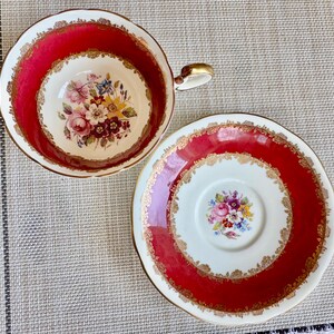 May include: A red and gold teacup and saucer set with a floral pattern. The cup and saucer have a white background with a red rim and gold trim. The floral pattern is in shades of pink, yellow, and blue.