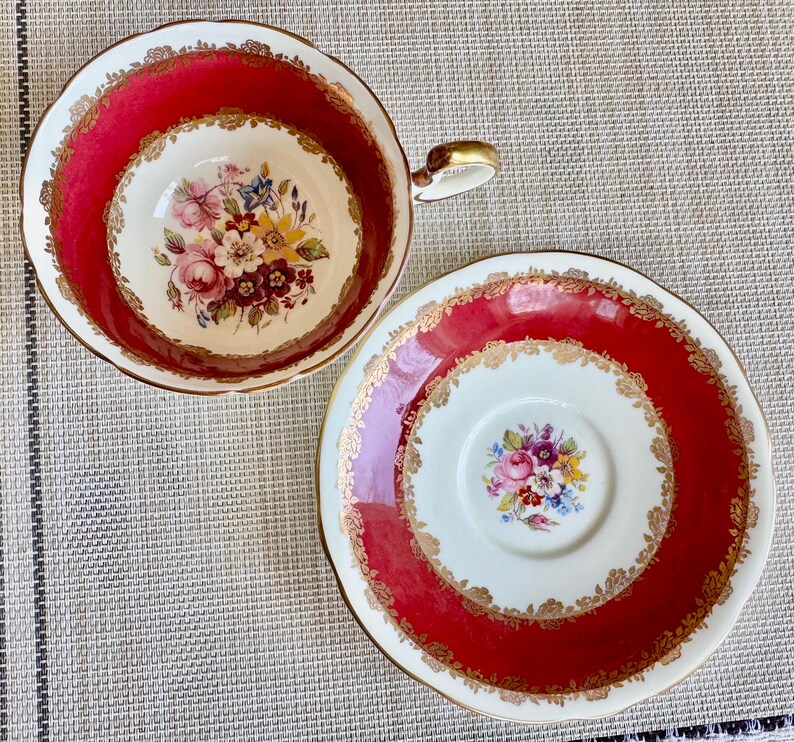 May include: A red and gold teacup and saucer set with a floral pattern. The cup and saucer have a white background with a red rim and gold trim. The floral pattern is in shades of pink, yellow, and blue.
