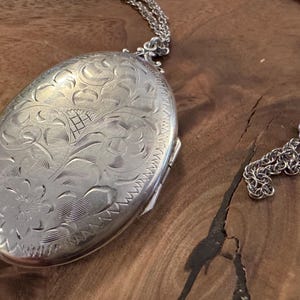 Puede incluir: Collar con colgante de medall&oacute;n ovalado plateado con intrincados grabados florales y de volutas. El medall&oacute;n cuelga de una delicada cadena plateada. El collar se muestra sobre una superficie de madera.