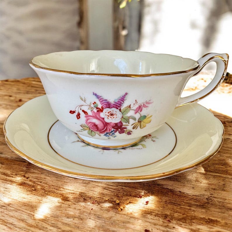 May include: A white teacup and saucer set with a floral design. The cup and saucer have a gold trim around the edge. The floral design features pink, purple, and yellow flowers.