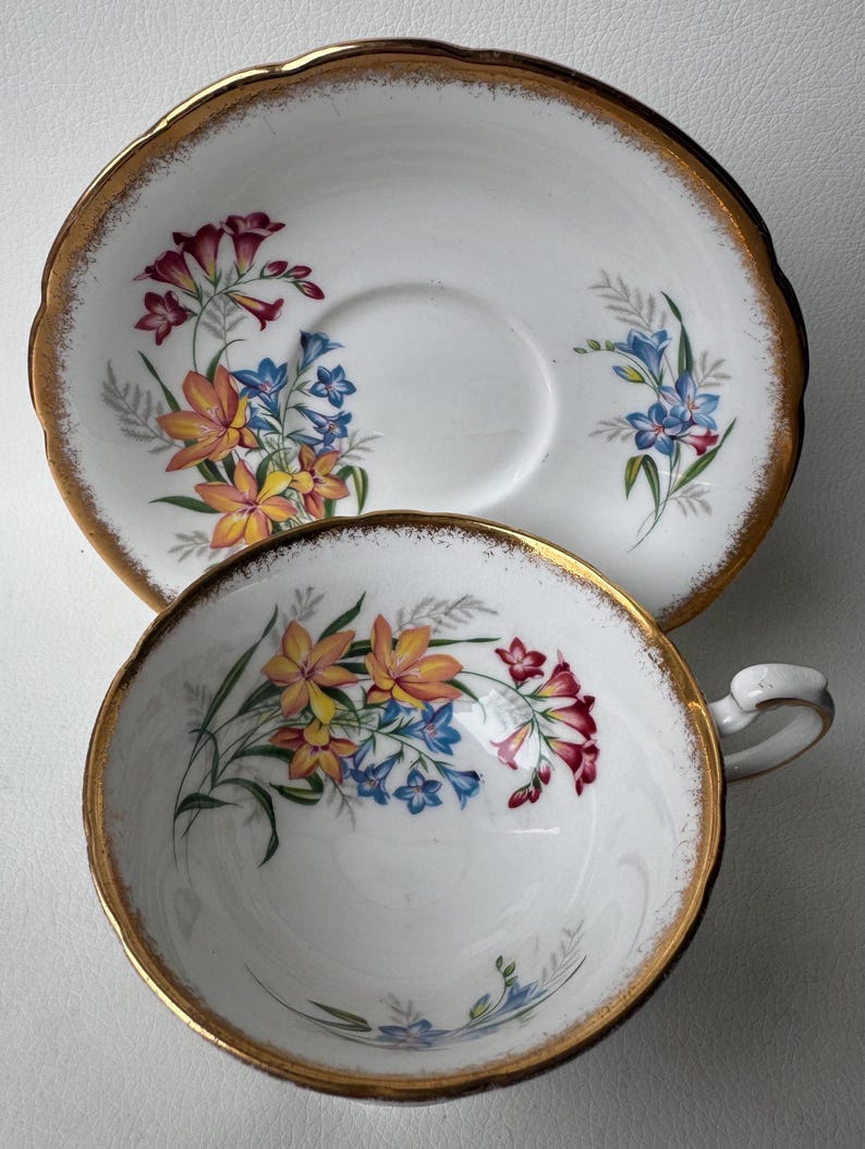 May include: A white teacup and saucer with a gold rim and a floral pattern of yellow, orange, blue, and red flowers.