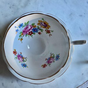 Puede incluir: Una taza y un platillo blancos con un dise&ntilde;o floral. La taza tiene un asa y un borde festoneado, y el platillo tiene un borde a juego. El patr&oacute;n floral incluye flores rosas, azules, amarillas y rojas con ribete dorado.