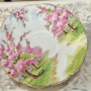 May include: A white teacup saucer with a scalloped edge, featuring a hand-painted scene of pink flowering trees and green grass. The saucer has a gold trim and is set on a silver tray.