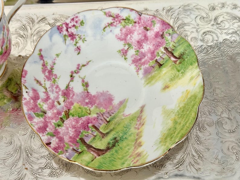 May include: A white teacup saucer with a scalloped edge, featuring a hand-painted scene of pink flowering trees and green grass. The saucer has a gold trim and is set on a silver tray.