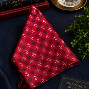 May include: A red pocket square featuring a repeating pattern of white anchors and dark blue ship wheels. The fabric has a subtle diagonal ribbed texture and is folded into a triangular shape, displaying a consistent nautical motif throughout.