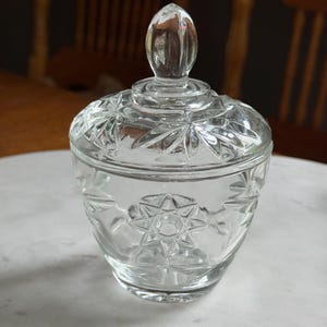 May include: A clear glass sugar bowl with a matching lid featuring a pointed finial. The exterior surface of the bowl and lid is decorated with a pressed starburst pattern and geometric facets that catch the light, creating a textured appearance.