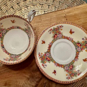 May include: A vintage teacup and saucer set with a floral and butterfly design. The cup and saucer are white with gold trim and feature a delicate floral pattern with colorful butterflies.