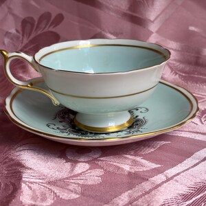 May include: A pale blue and white porcelain teacup and saucer set featuring gold trim along the scalloped edges and base. The saucer displays a dark gray ornate floral pattern, while the cup has a smooth interior and a gold-accented handle.