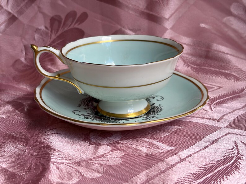 May include: A pale blue and white porcelain teacup and saucer set featuring gold trim along the scalloped edges and base. The saucer displays a dark gray ornate floral pattern, while the cup has a smooth interior and a gold-accented handle.