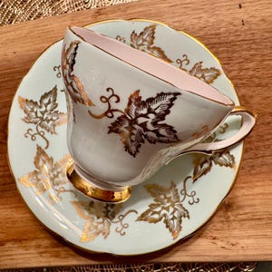May include: A vintage teacup and saucer set with a delicate floral design. The cup and saucer are white with a light blue background and gold accents. The floral design features leaves and vines.