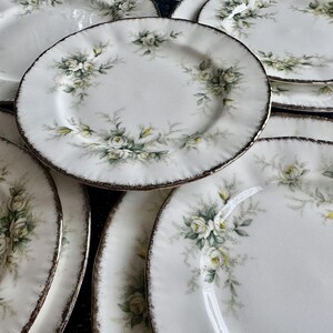 May include: A set of white ceramic dinner plates with a floral design featuring white roses and green leaves. The plates have a slightly scalloped edge and a gold rim.