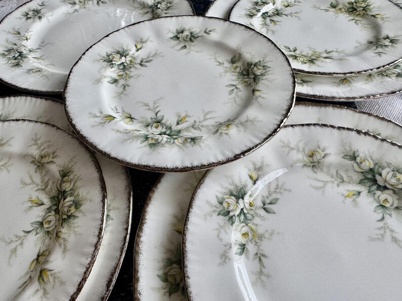 May include: A set of white ceramic dinner plates with a floral design featuring white roses and green leaves. The plates have a slightly scalloped edge and a gold rim.