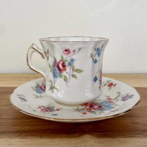 May include: A white teacup and saucer set with a floral pattern in pink, blue, and purple. The cup has a gold handle and rim. The saucer has a scalloped edge and gold trim. The set is designed for tea or coffee.