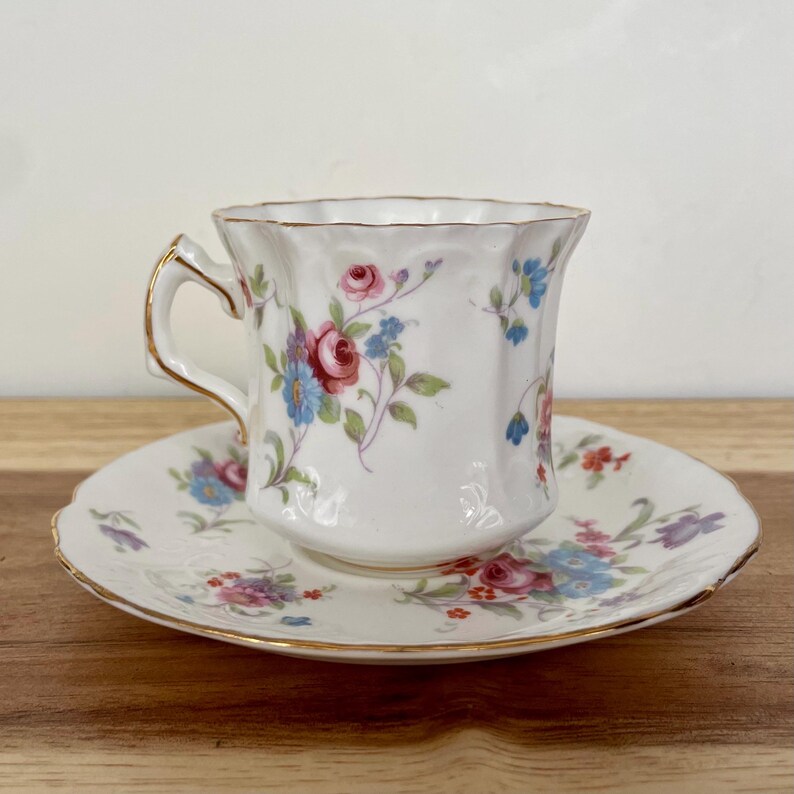May include: A white teacup and saucer set with a floral pattern in pink, blue, and purple. The cup has a gold handle and rim. The saucer has a scalloped edge and gold trim. The set is designed for tea or coffee.