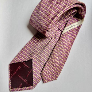 Puede incluir: Una corbata Salvatore Ferragamo con un patr&oacute;n repetitivo de peque&ntilde;os motivos en forma de arco de color claro sobre un fondo morado. La corbata incluye una etiqueta de marca y un forro morado oscuro con el logotipo de Salvatore Ferragamo impreso.