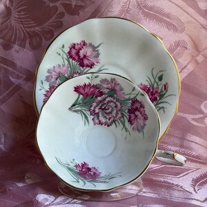 May include: A white porcelain teacup and saucer set featuring a delicate floral pattern of magenta carnations with green stems and leaves. Both the cup and the saucer have scalloped edges trimmed with a thin gold band, and the cup has a small handle.