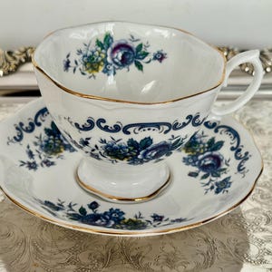以下が含まれることがあります： 白のティーカップとソーサーのセット。青、緑、紫の色合いの花柄模様。カップとソーサーの縁はスカラップで、金色のトリムが施されています。クラシックなティーセット。