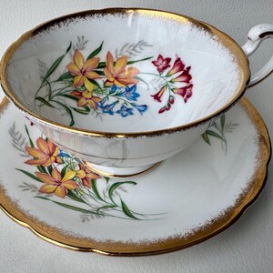 May include: A white teacup and saucer set with a gold rim and floral pattern. The flowers are yellow, orange, pink, and blue.