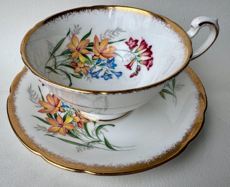 May include: A white teacup and saucer set with a gold rim and floral pattern. The flowers are yellow, orange, pink, and blue.