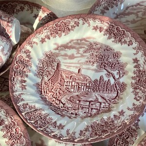 Pu&ograve; includere: Una collezione di piatti in ceramica bianca e bordeaux. I piatti e le tazze presentano una scena pastorale dettagliata con una casa, alberi e un ponte. I bordi sono decorati con motivi floreali e di vite. I piatti sono disposti su una superficie.