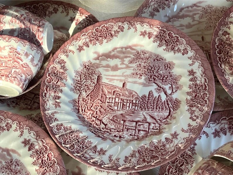 Pu&ograve; includere: Una collezione di piatti in ceramica bianca e bordeaux. I piatti e le tazze presentano una scena pastorale dettagliata con una casa, alberi e un ponte. I bordi sono decorati con motivi floreali e di vite. I piatti sono disposti su una superficie.