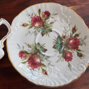 May include: A white porcelain teacup and saucer with a gold rim. The saucer features a floral design with red roses and green leaves.