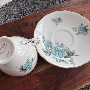 May include: A white teacup and saucer set with a blue floral pattern. The saucer has a gold rim and the teacup has the words "Royal China Colclough Made in England" on the bottom.