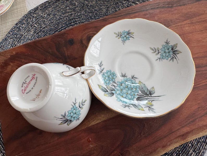 May include: A white teacup and saucer set with a blue floral pattern. The saucer has a gold rim and the teacup has the words "Royal China Colclough Made in England" on the bottom.