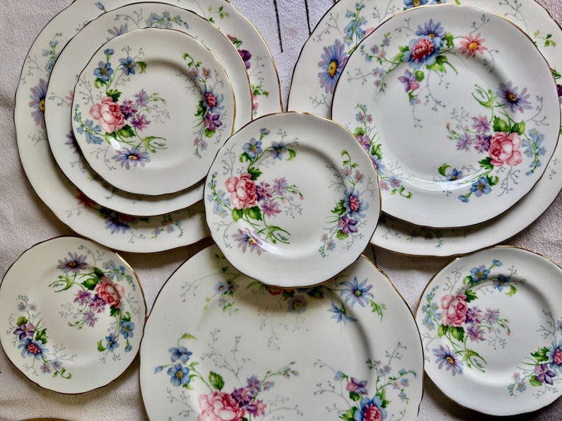May include: A selection of white porcelain plates with a floral pattern, including pink roses, purple and blue flowers, and gold edging. The set comprises various plate sizes, from small saucers to large dinner plates, arranged on a white surface.